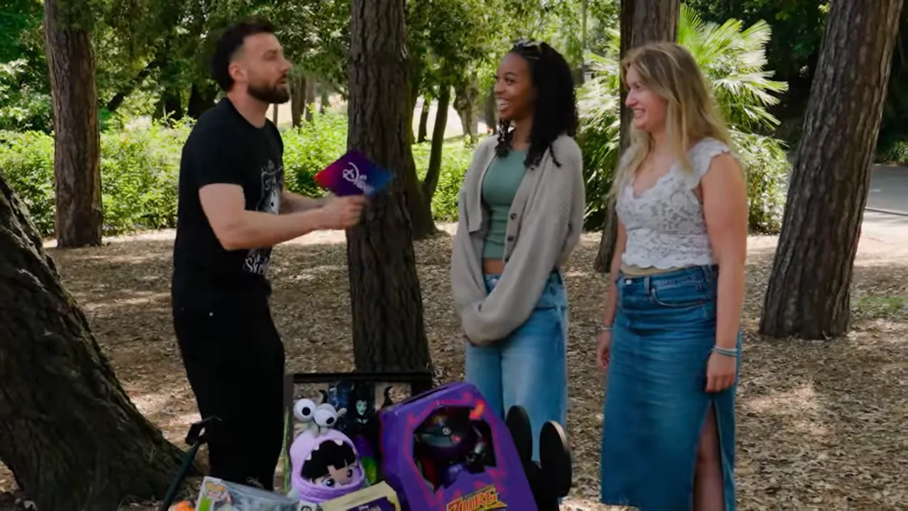 Host talking to two contestants beside a cart of Disney-themed prizes in a park.