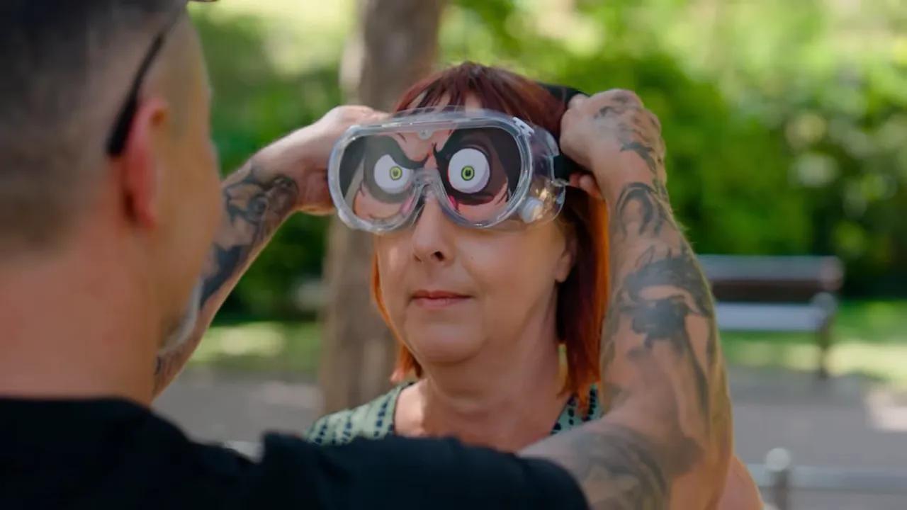 Host placing oversized novelty goggles on a participant during a street game in a park.