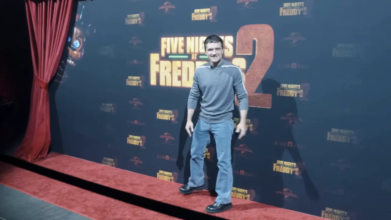 Actor Josh Hutcherson standing on a red carpet in front of a Five Nights at Freddy's 2 backdrop with an animatronic peeking from the curtain.