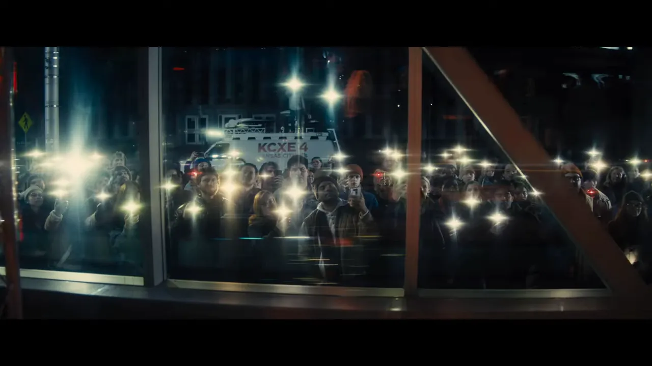 Crowd gathered at night holding up phones with bright lights and a news van parked behind them