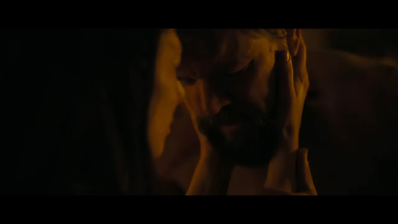 Intimate close-up of a bearded man being cradled by another's hands in warm low light