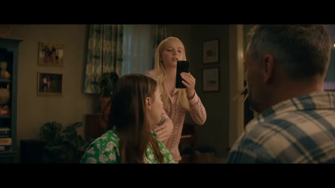 Father at table with two daughters, one holding up a phone in a family living room
