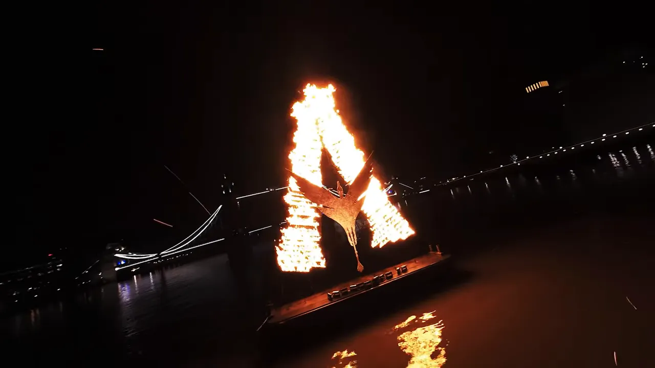 Nighttime drone view of a burning 'A' installation on a barge on the River Thames with an illuminated bridge and city lights behind