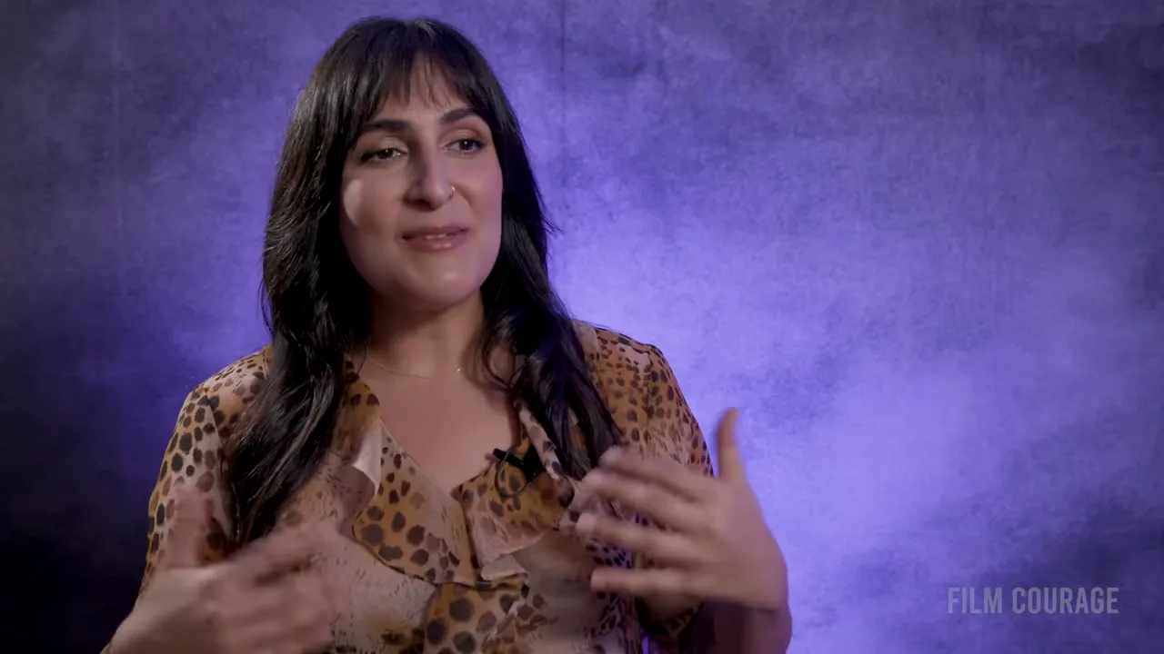 Interview subject smiling and gesturing with both hands against a textured purple backdrop, mid-explanation.