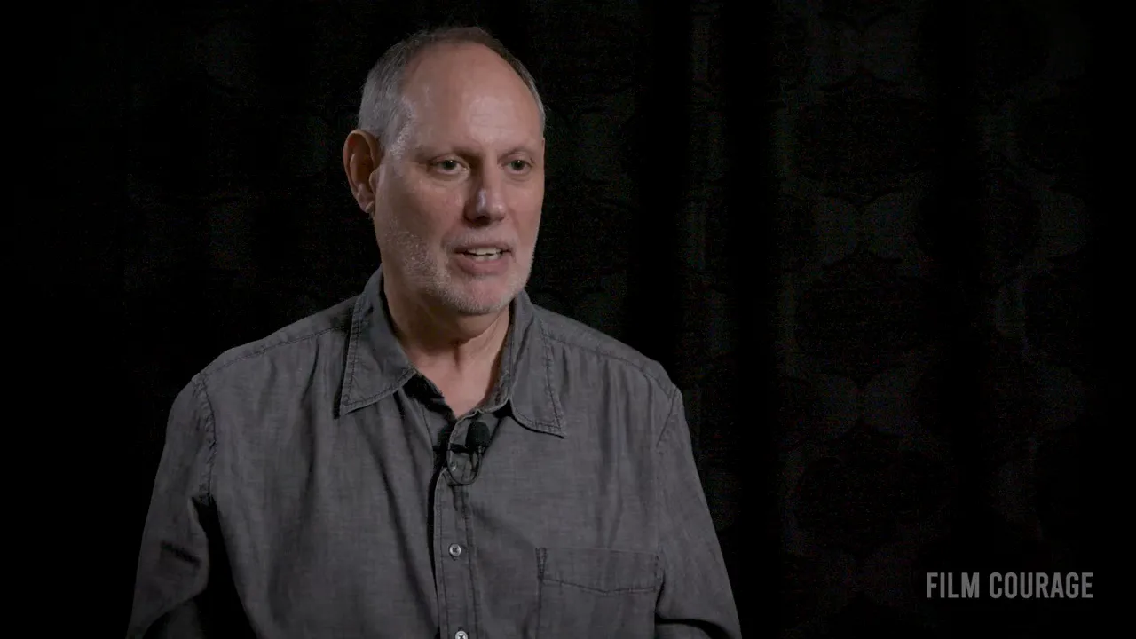 Interviewee speaking in a gray shirt against a dark backdrop, close-up.