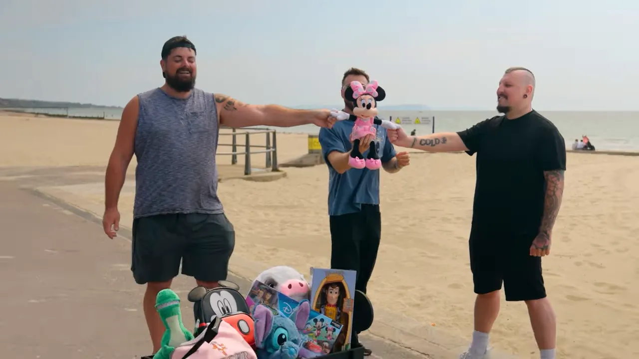 Two contestants standing on a beach each holding one hand of a Minnie Mouse plush, with a wagon of Disney prize toys in the foreground.
