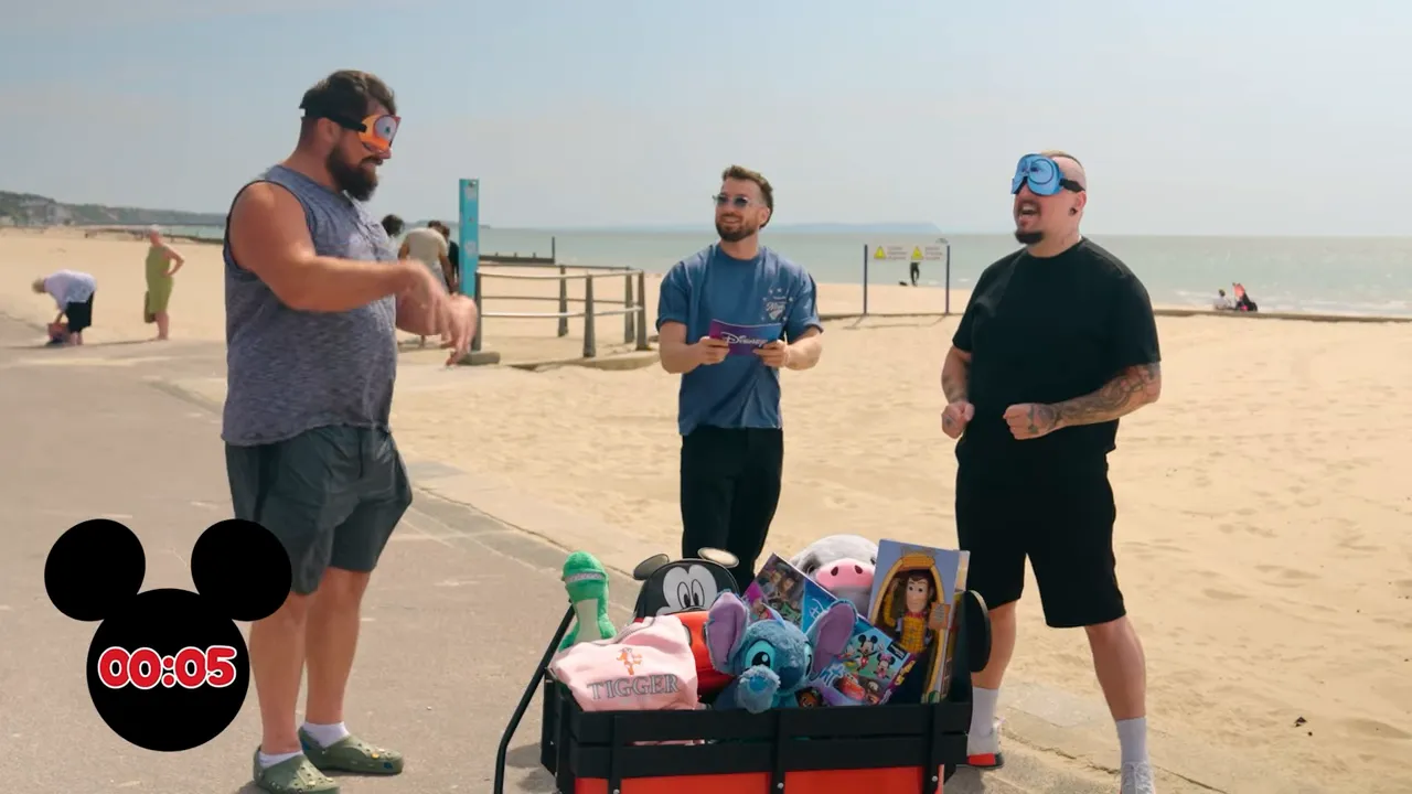 Two blindfolded contestants laughing and gesturing with the host between them and a wagon of Disney plush toys on the beach