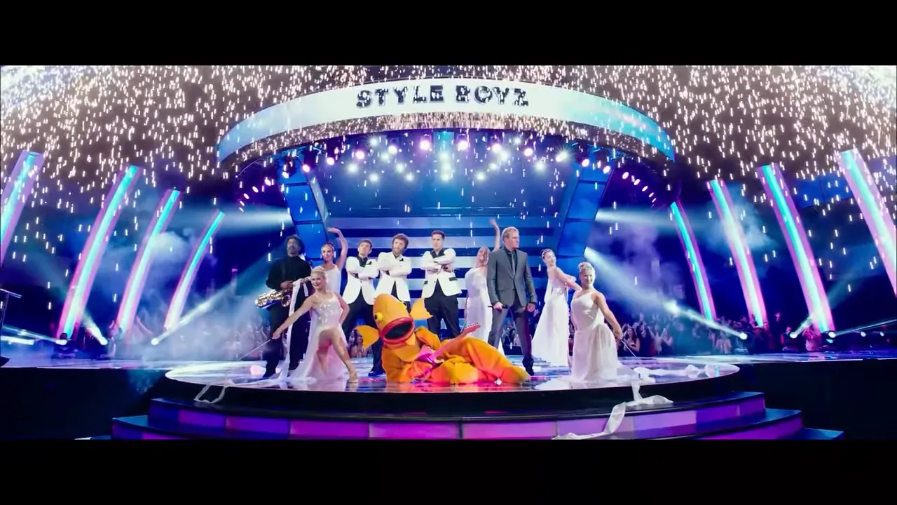 Centered wide shot of the Style Boyz and dancers onstage with a large fish costume under a 'STYLE BOYZ' marquee as sparks fall and bright lights illuminate the set.