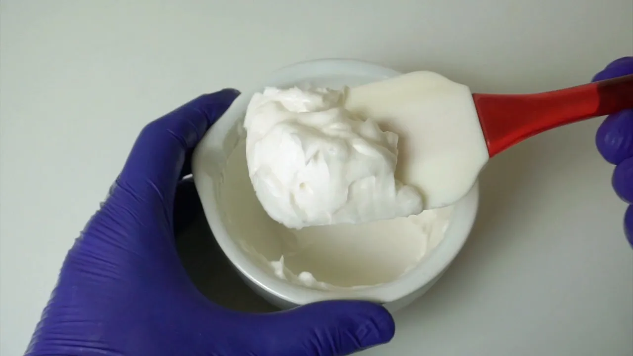 Espátula roja levantando una porción de emulsión blanca homogénea de un recipiente sostenido con guante morado