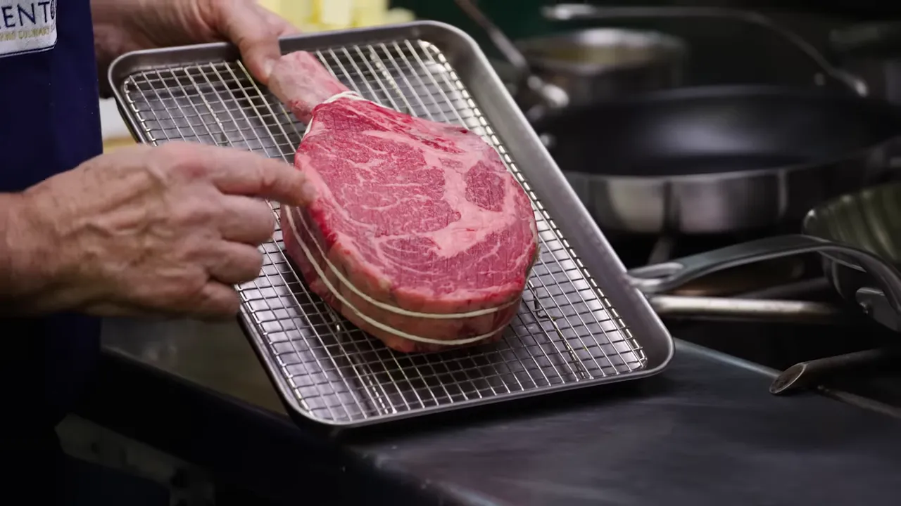 Bone-in ribeye steak secured with string resting on a wire rack in a stainless-steel kitchen