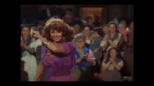 Woman in a purple dress dancing in front of a cheering audience in a color film scene.
