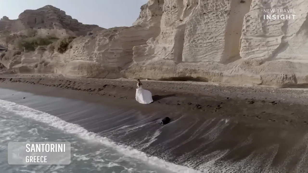 Person in white clothing standing near the black sand and waves at Santorini, Greece
