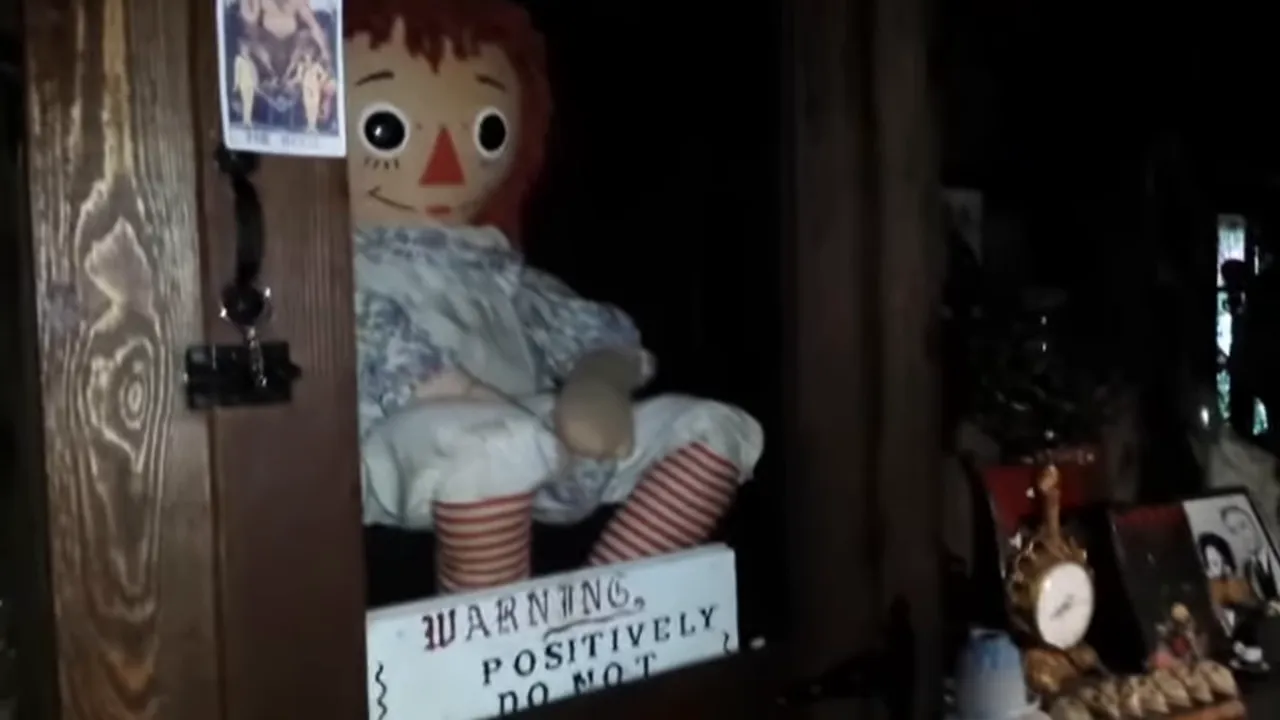 Raggedy Ann doll seated in a wooden display case with a warning sign below