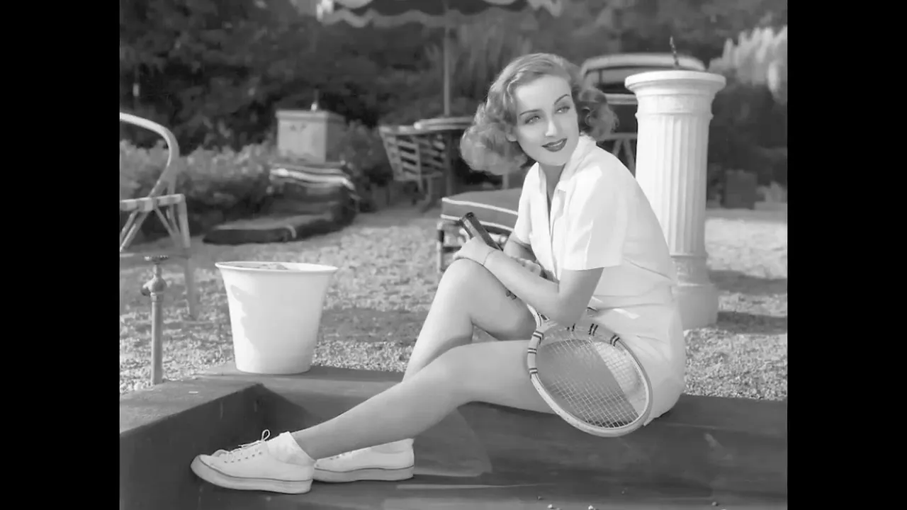 Black-and-white photo of Carole Lombard seated outdoors holding a tennis racquet, in casual tennis attire