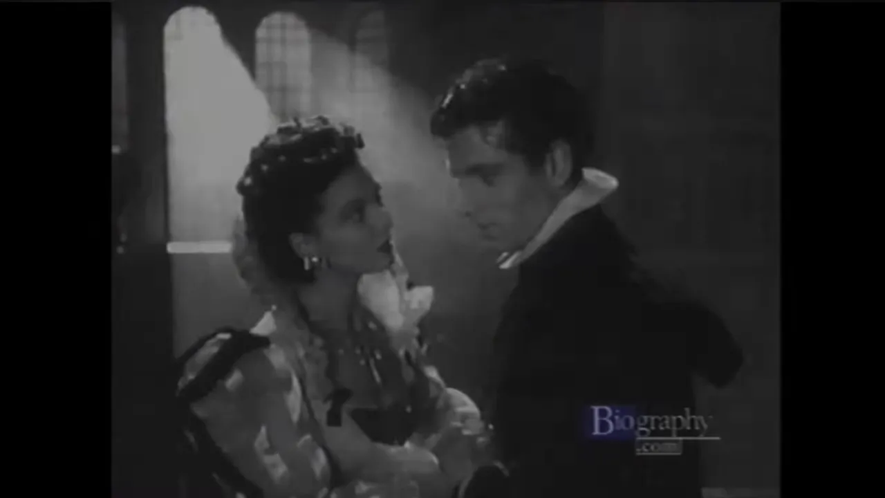 Black-and-white film still of a man and woman in period costume facing each other closely in a tense romantic scene.