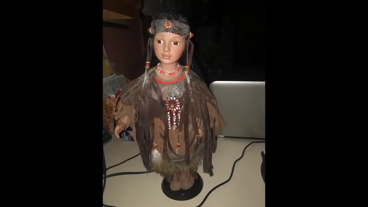 clear front view of a vintage doll dressed in feathers and beads on a desk with a laptop in the background