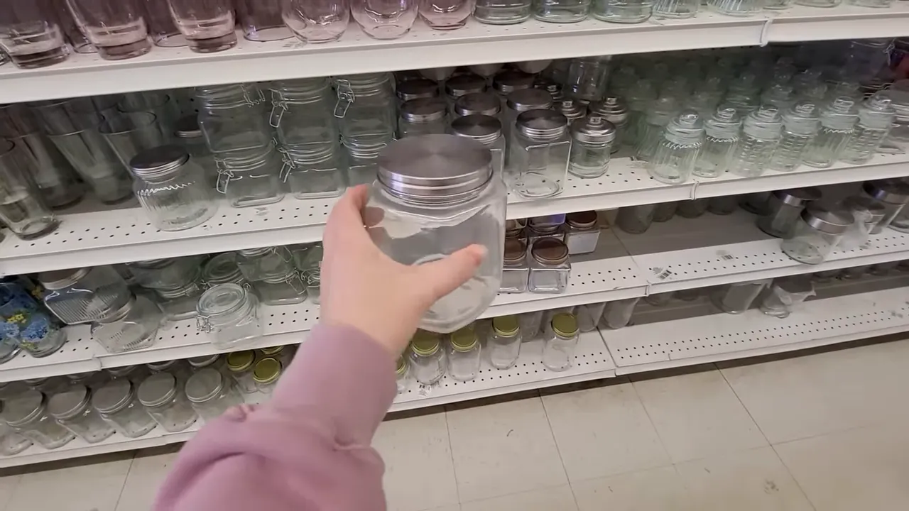 Hand holding a clear glass jar with a silver metal lid in front of store shelves of jars