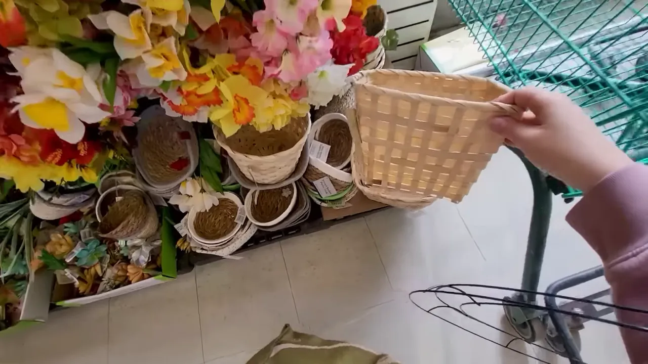 Hand holding a light woven basket in a craft store aisle next to faux florals and a wire display rack