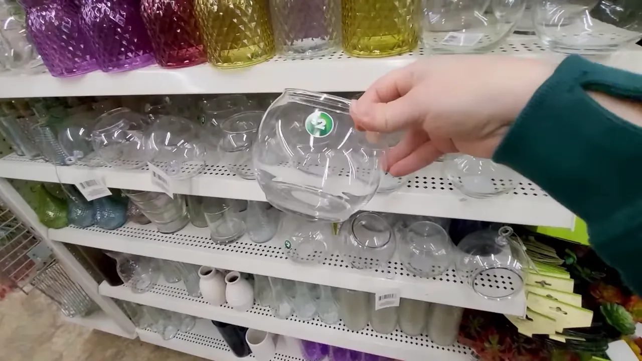 Hand holding a clear round glass vase in front of store shelving with multiple glass vases