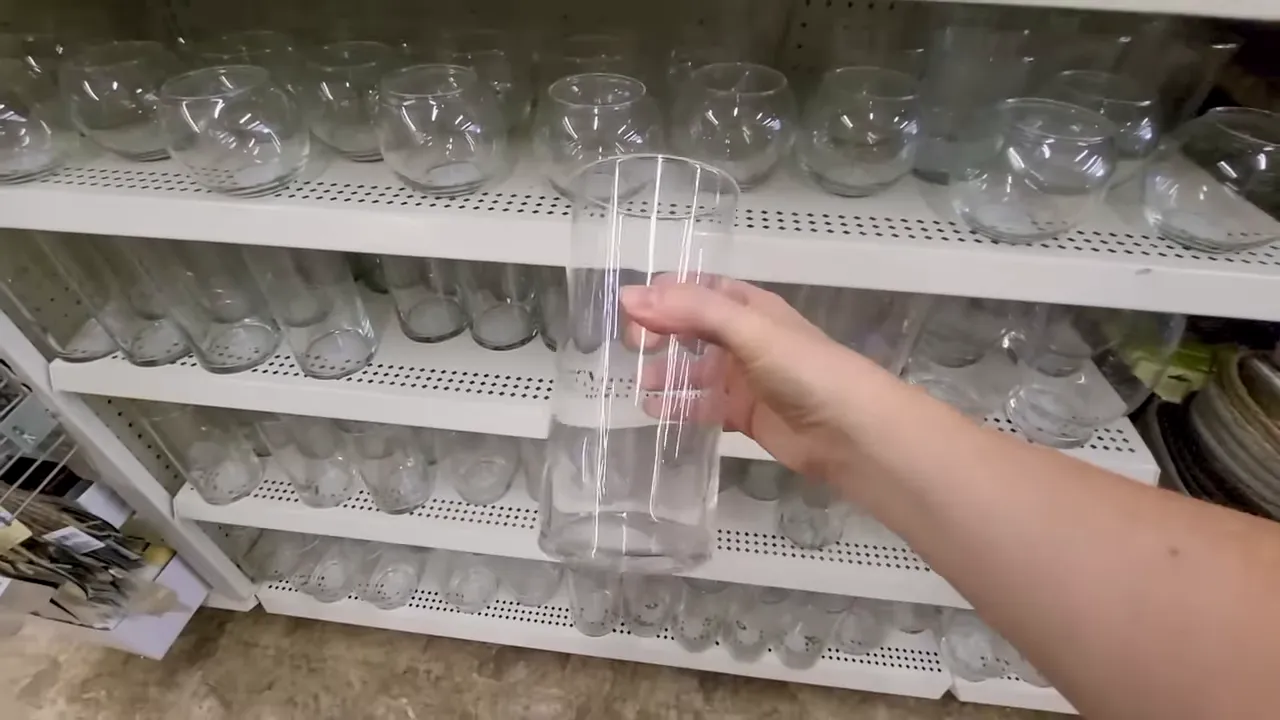 Hand holding a clear tall glass vase in front of store shelves filled with glass vases