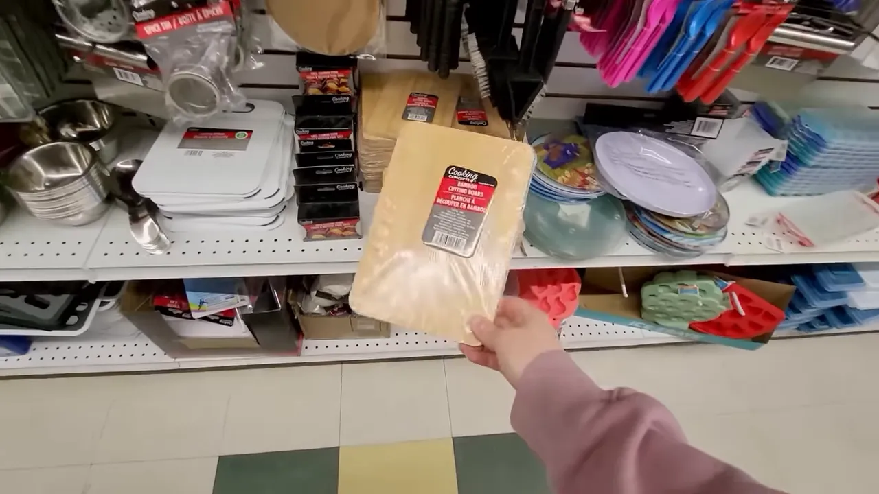 Hand holding a small packaged wooden cutting board in a store aisle, showing size and label