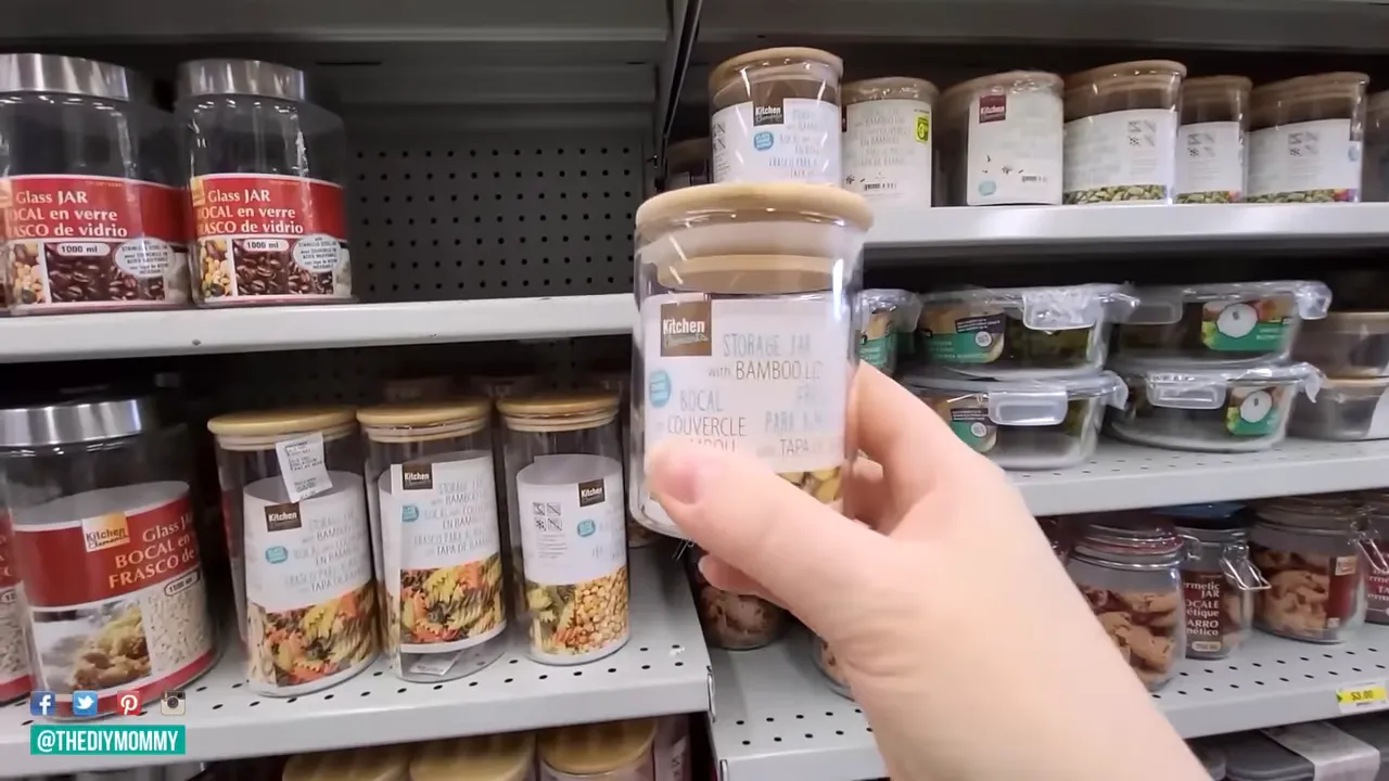 Hand holding a small clear glass storage jar with a bamboo lid in front of store shelving filled with similar jars.