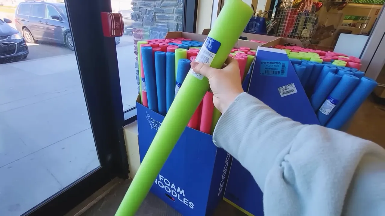 Hand holding a green foam pool noodle in front of bins of colorful foam noodles inside a craft store
