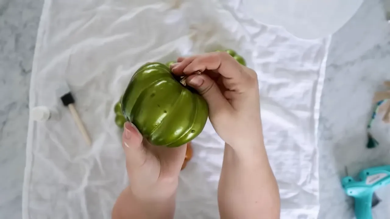 Overhead shot of hands holding a small green faux pumpkin on a cloth with craft tools nearby