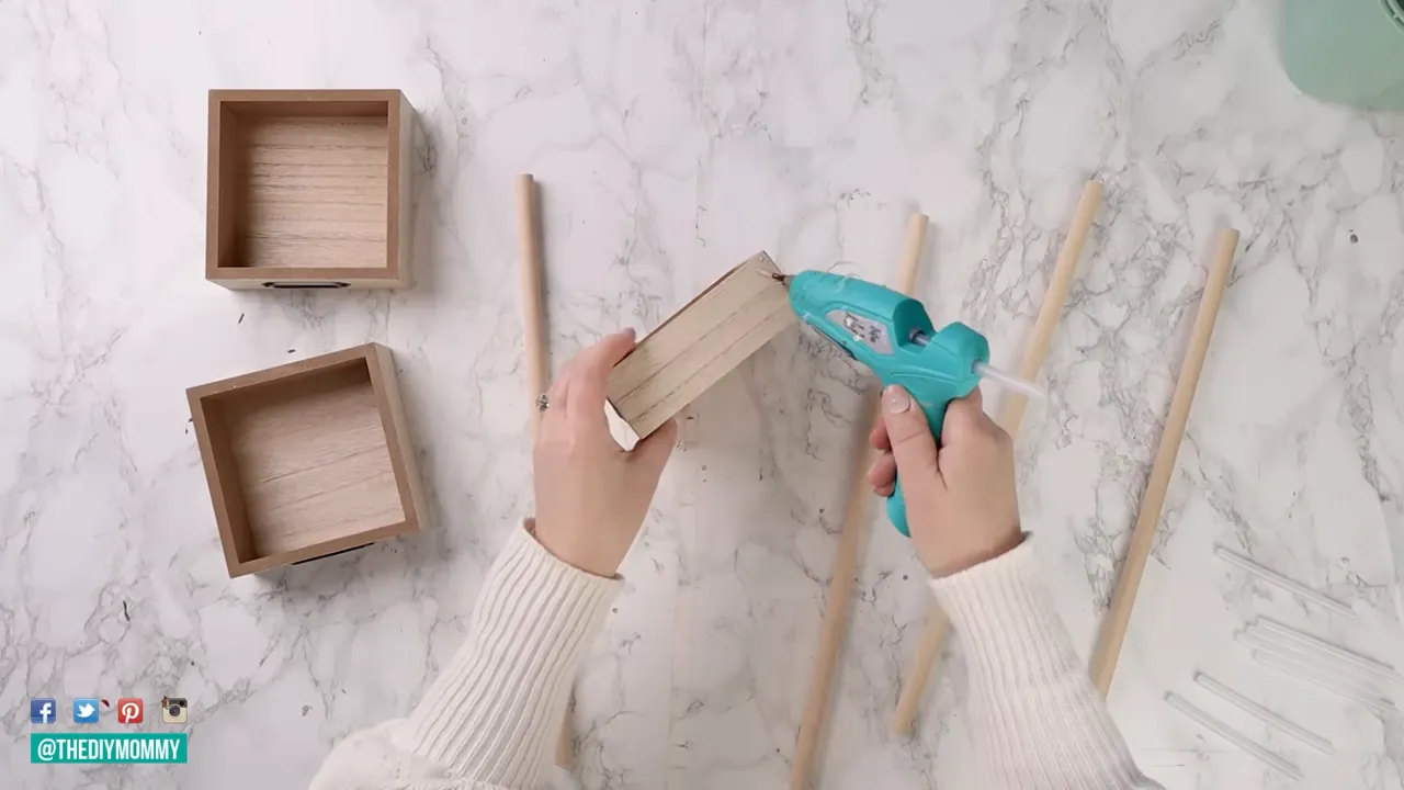 Overhead view of hands applying hot glue to the edge of a small wooden craft box with wooden dowels and glue sticks nearby, ready to build a tiered organizer.