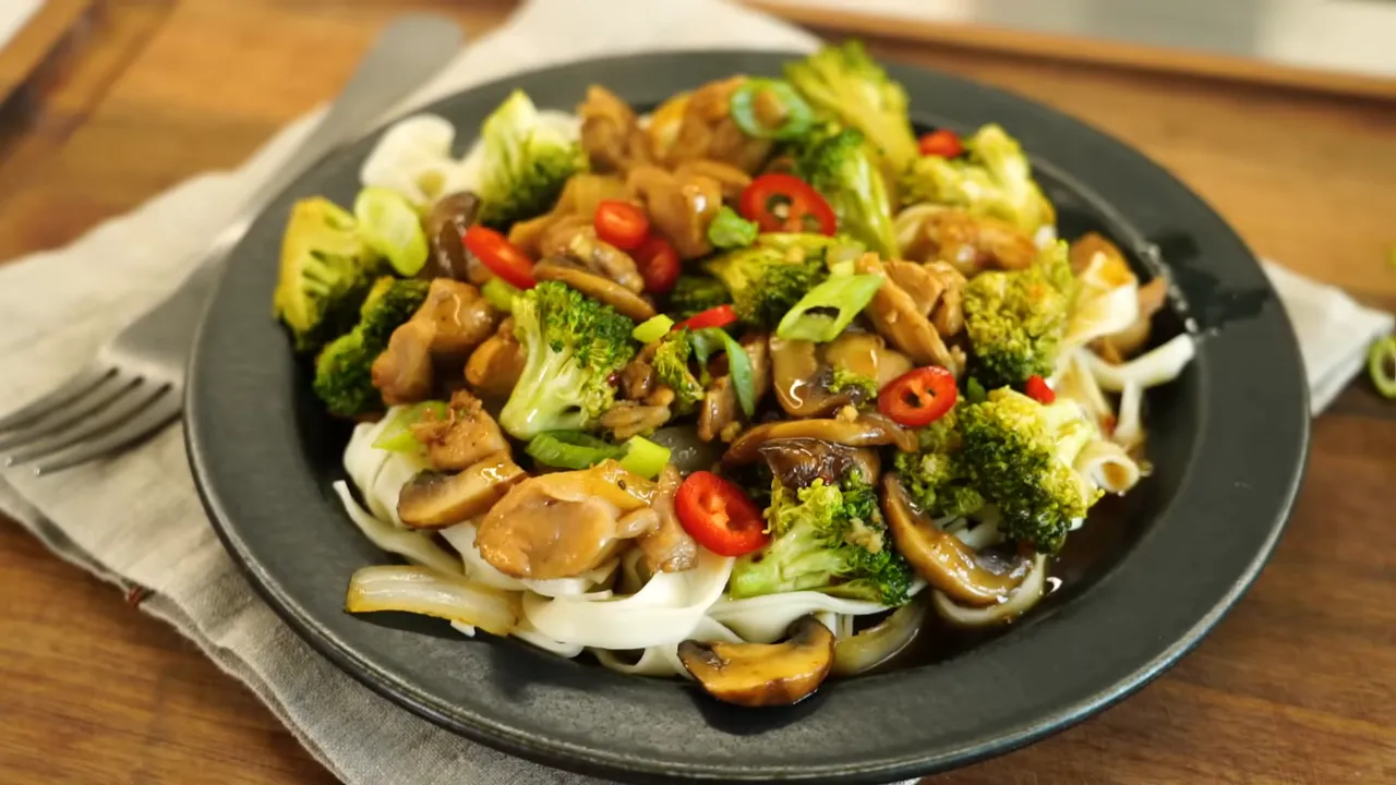 Chicken broccoli mushroom and onion stir-fry served over noodles on a dark plate