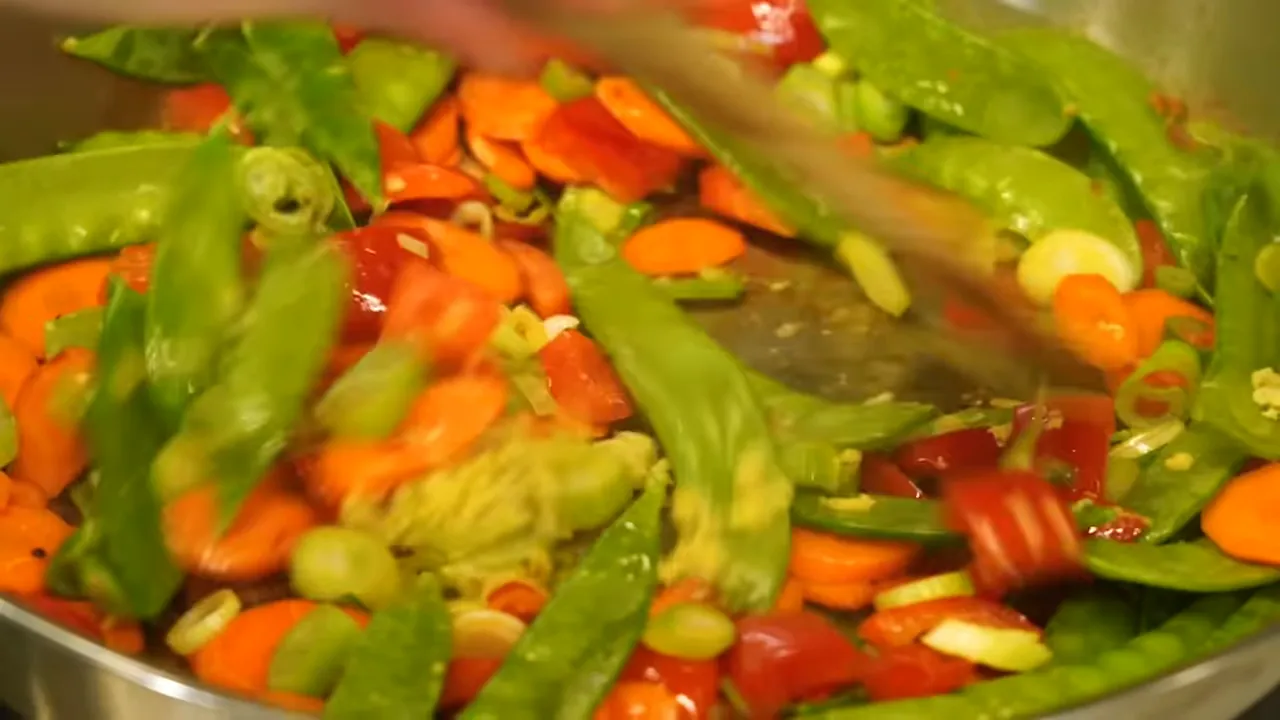 Brown stir-fry sauce being poured over carrots bell peppers snow peas and green onions in a skillet