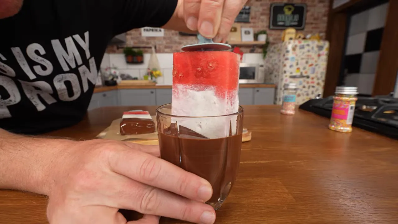 Frozen watermelon wedge with white yogurt layer being dipped into melted chocolate in a glass