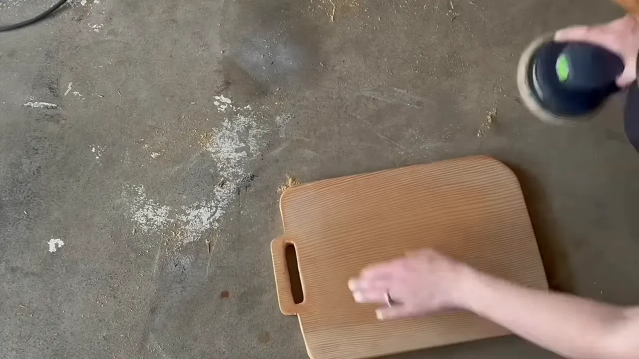Top-down view of a rectangular wooden cutting board on a concrete floor with hands and an orbital sander preparing the surface