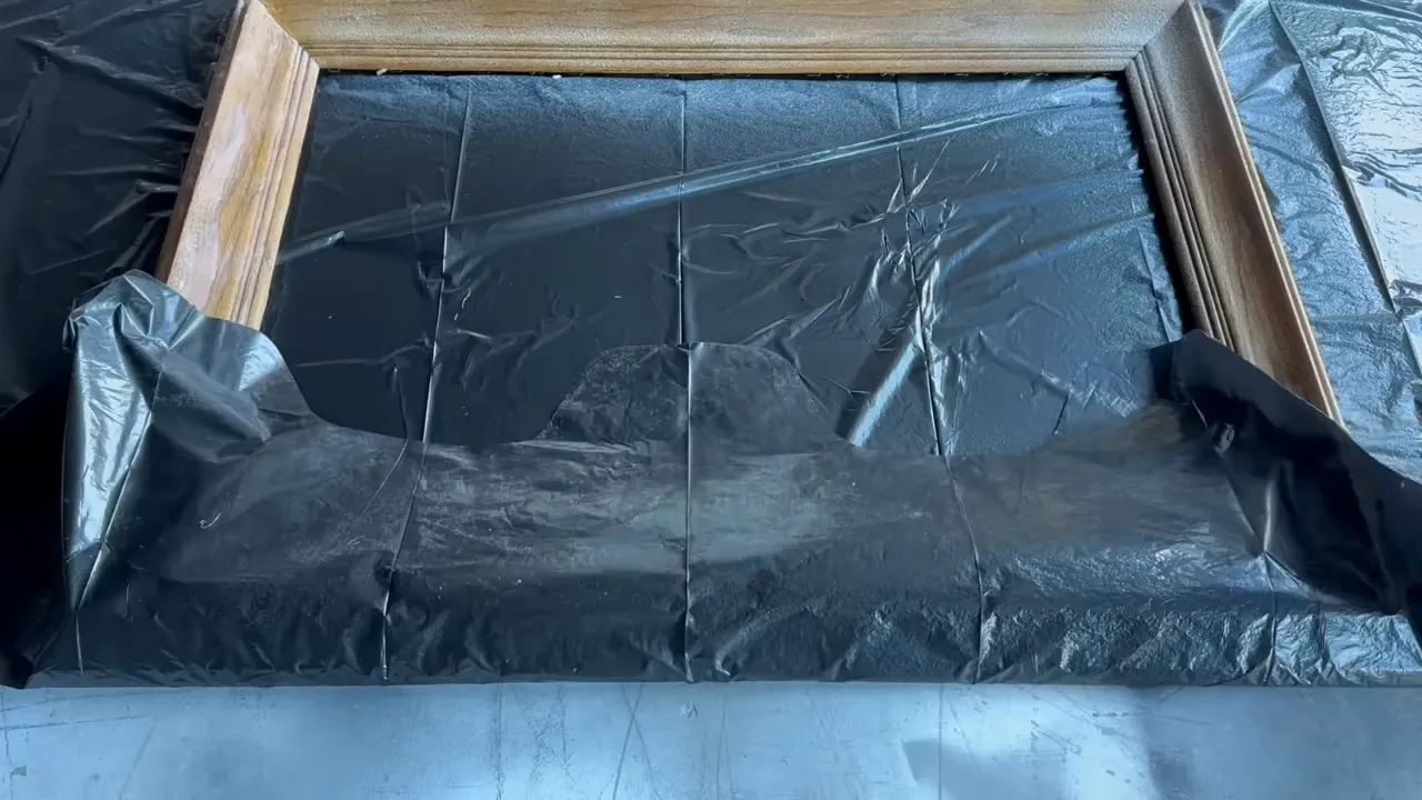 Wooden frame partially covered with black plastic sheeting, showing how the piece is wrapped to retain chemical stripper moisture
