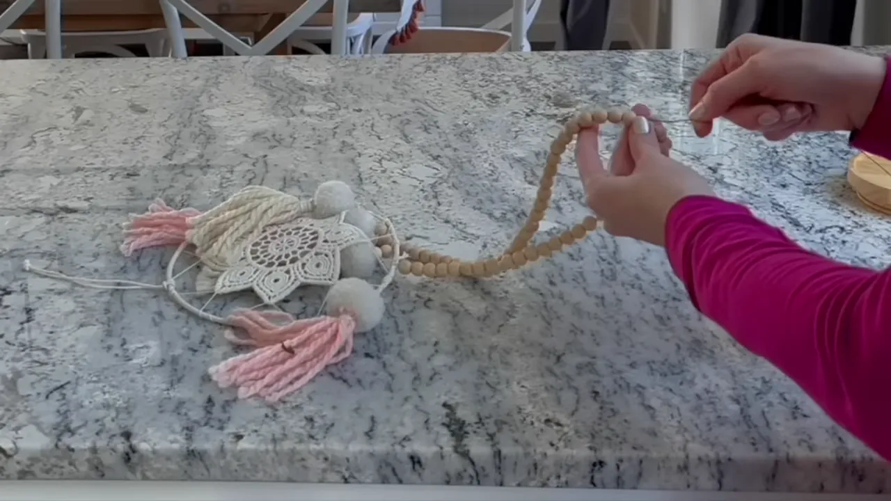 Hands threading a strand of wooden beads onto cord with a needle over a marble countertop