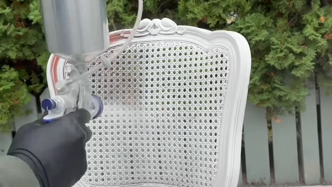 Gloved hand holding a spray gun applying white paint to a caned chair back outdoors.