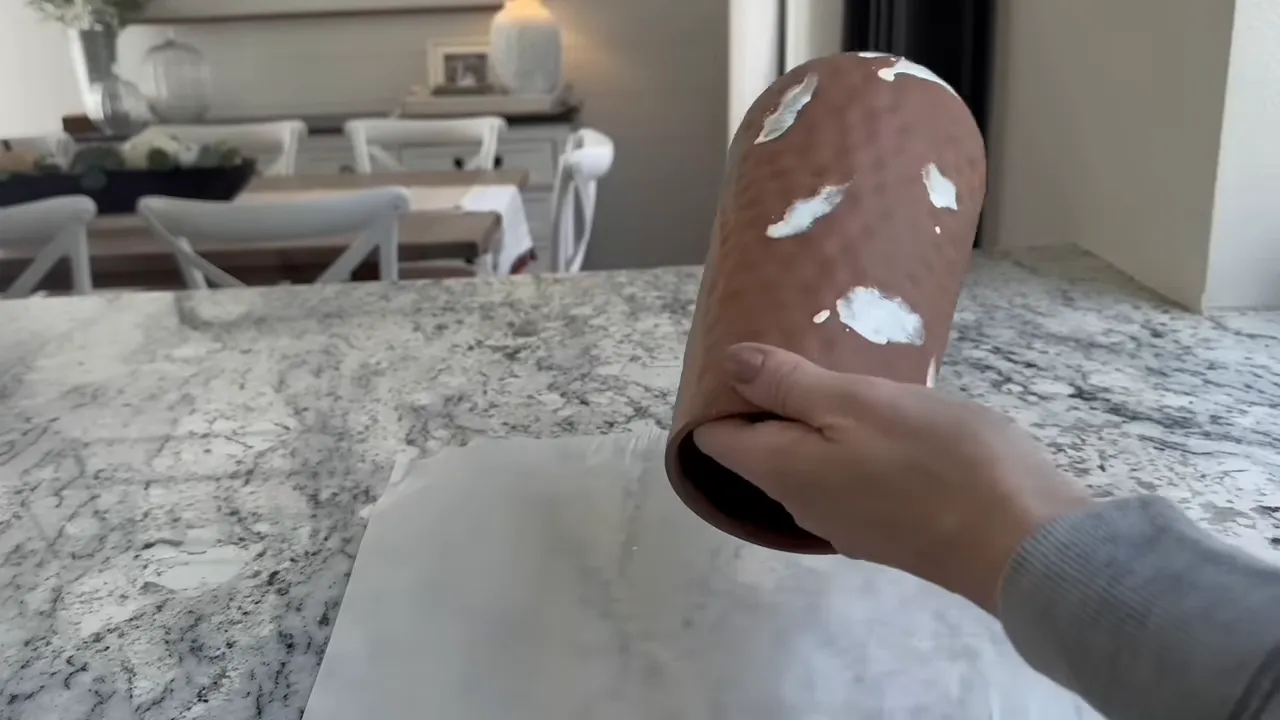 Hand holding a textured ceramic vase showing white paint drips and spots against a kitchen background
