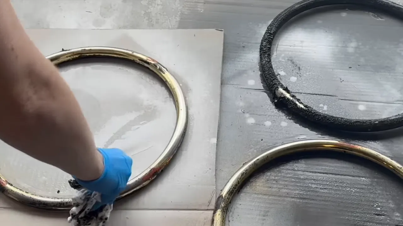 Gloved hand scrubbing brass-plated metal rings on a work surface with a rag