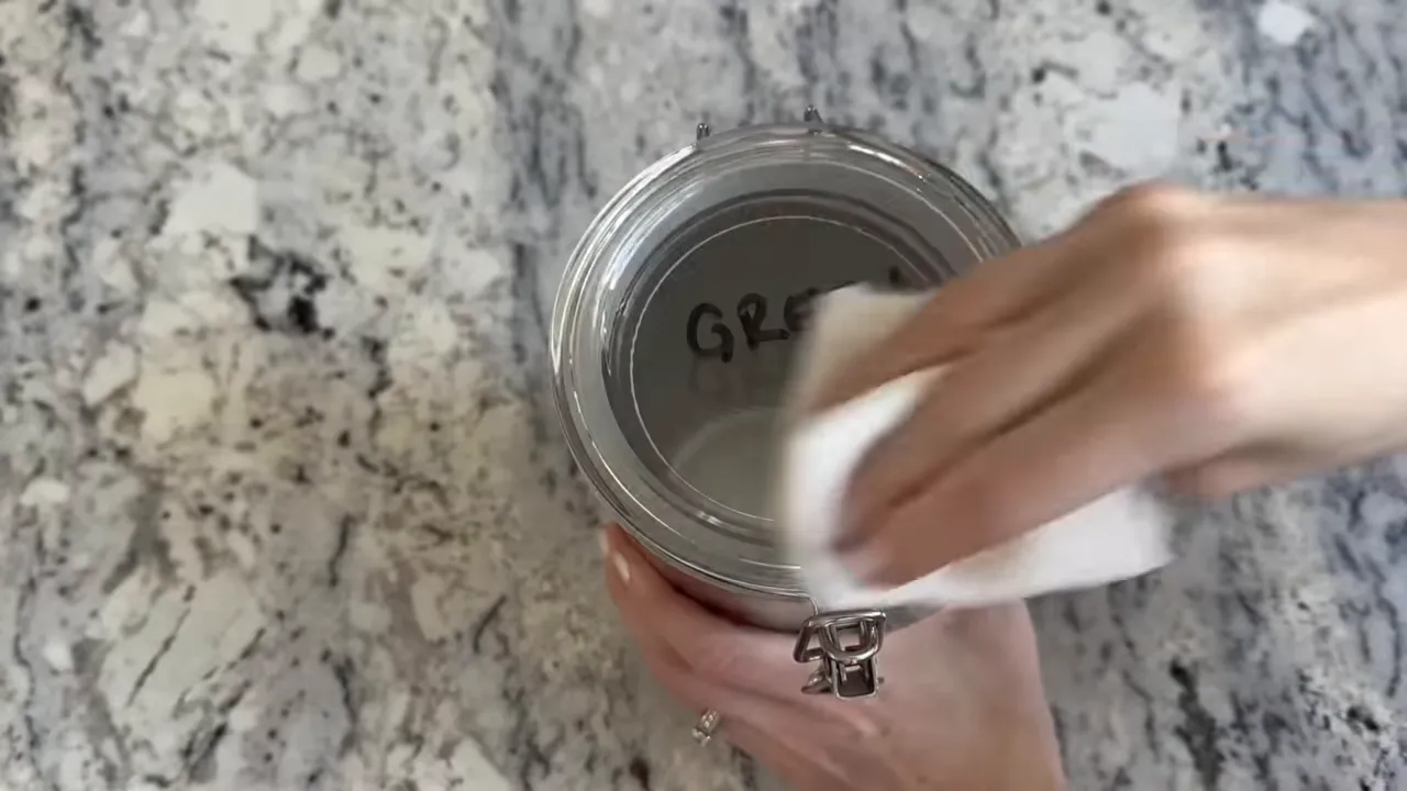 hands wiping a glass jar labeled 'GREEN' with a white cloth on a marble countertop to remove black marker