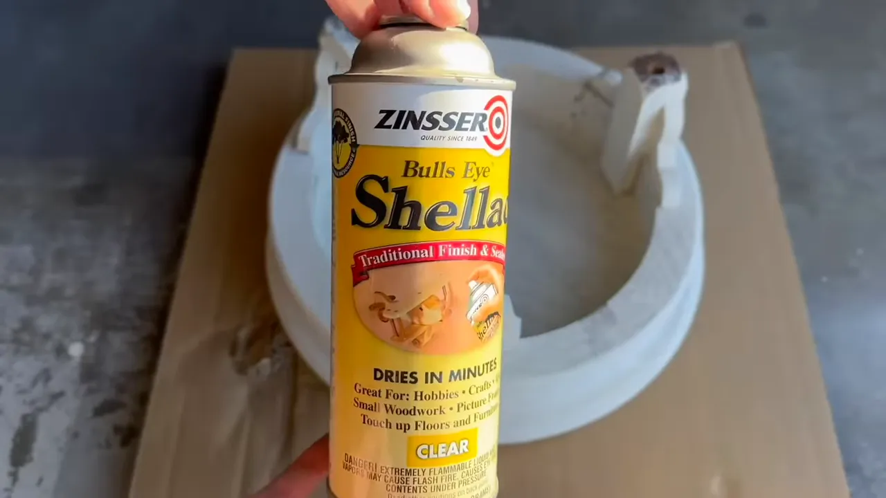 Hand holding a can of Zinsser Bulls Eye Shellac spray in front of a small painted wooden stool on cardboard; label clearly visible