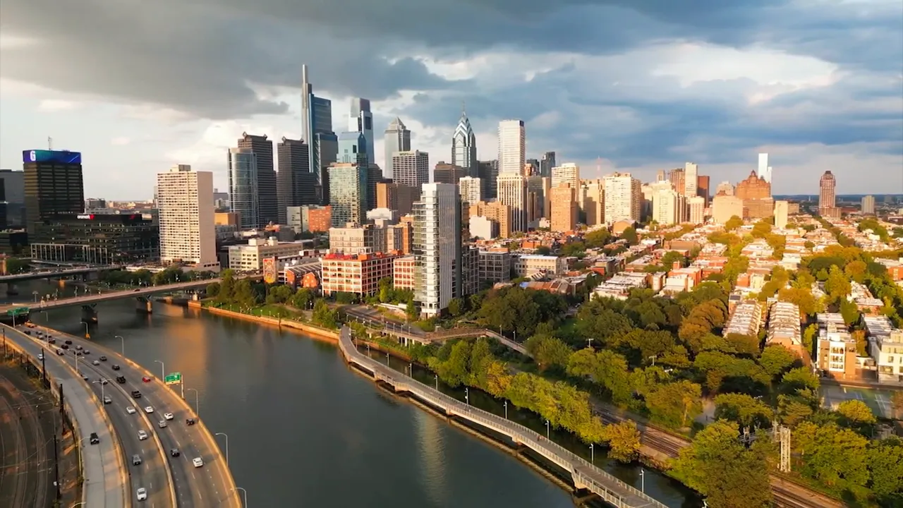 Golden-hour aerial view of Philadelphia skyline, riverwalk, highway and tree-lined neighborhoods