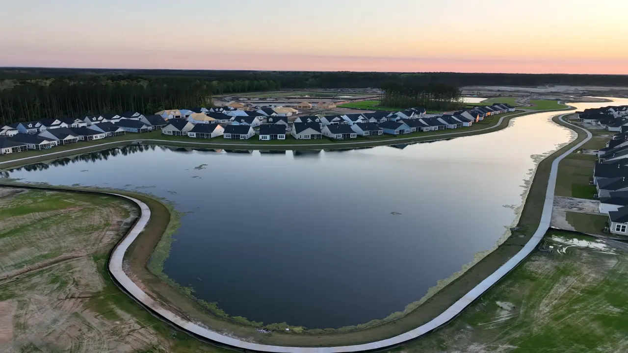 Aerial drone view of a large neighborhood pond with a surrounding walking path and rows of homes at sunset.