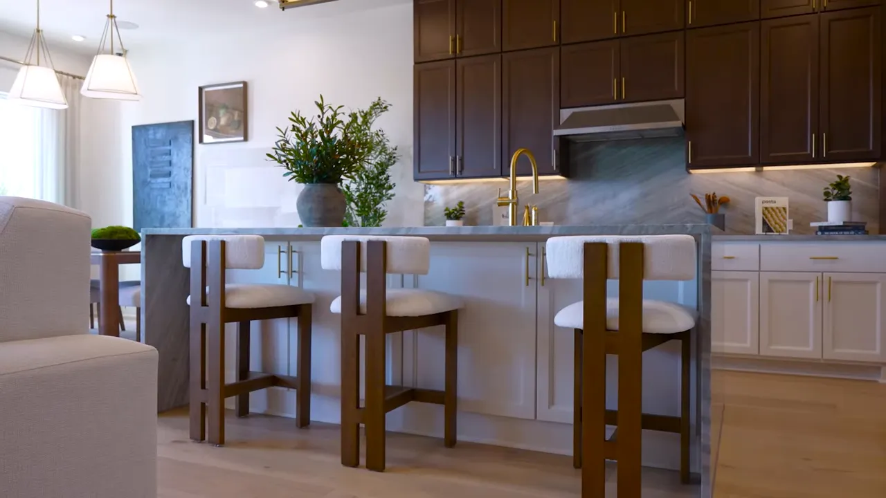 Open-plan kitchen island with centered gold faucet, three bar stools, dark upper cabinets and living area in the background