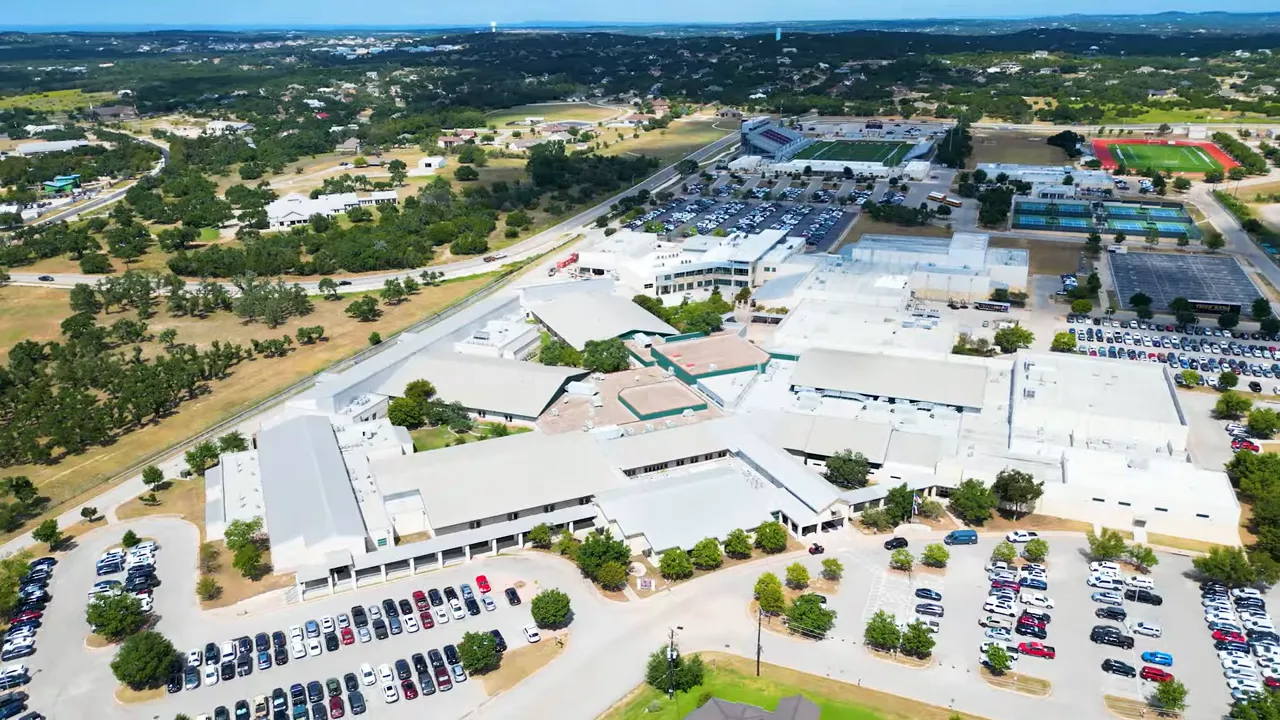 High-angle drone photo of a school campus with parking lots, football stadium, tennis courts and surrounding Hill Country landscape.