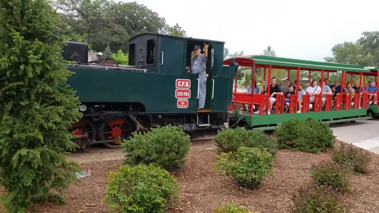 Small green locomotive pulling open red-and-green passenger cars through landscaped park grounds with riders visible.