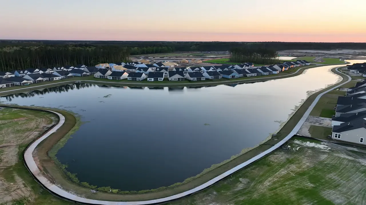 Expansive aerial of a large man‑made lake with a winding walking path and houses along the shoreline at sunset.