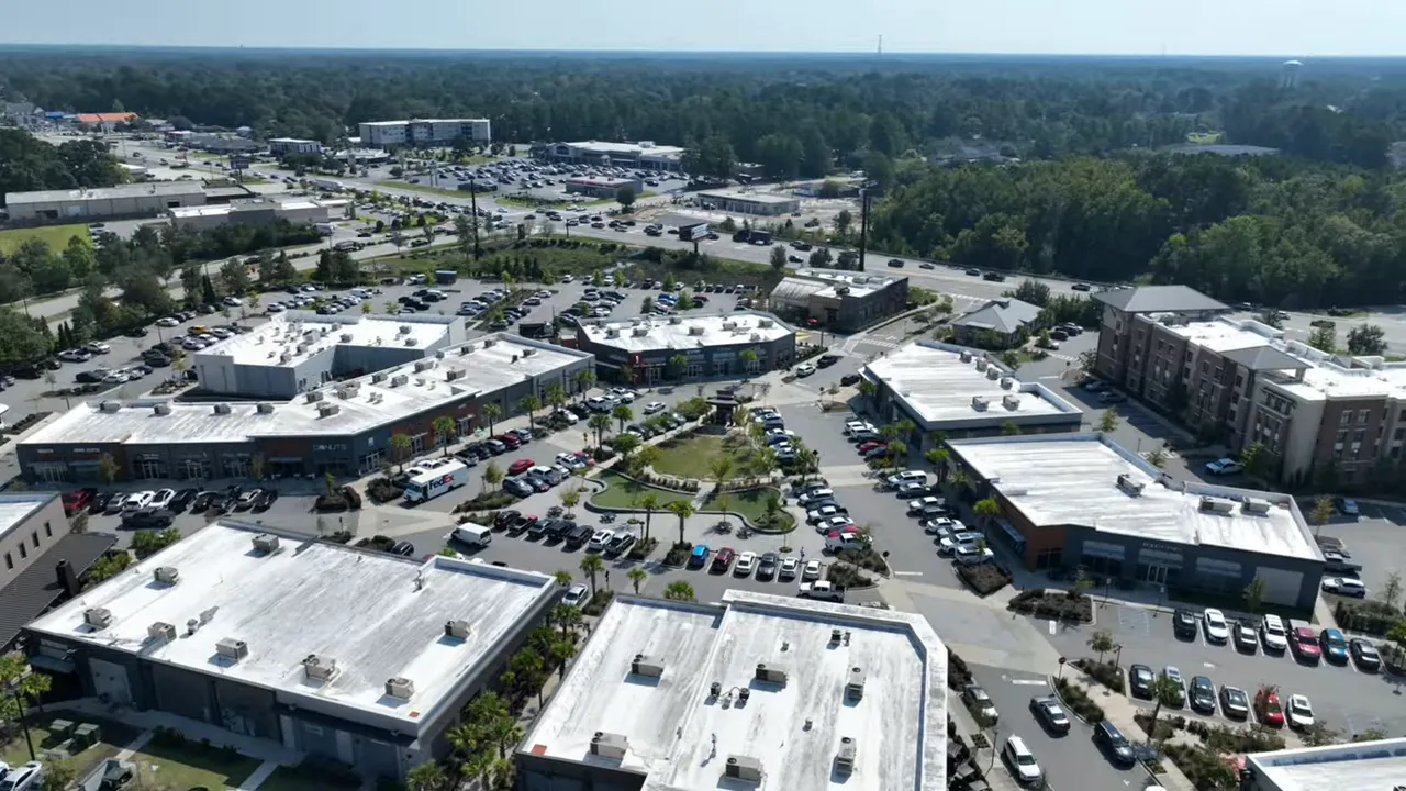 High-resolution drone view of a town center plaza with retail buildings, parked cars and landscaped green space.