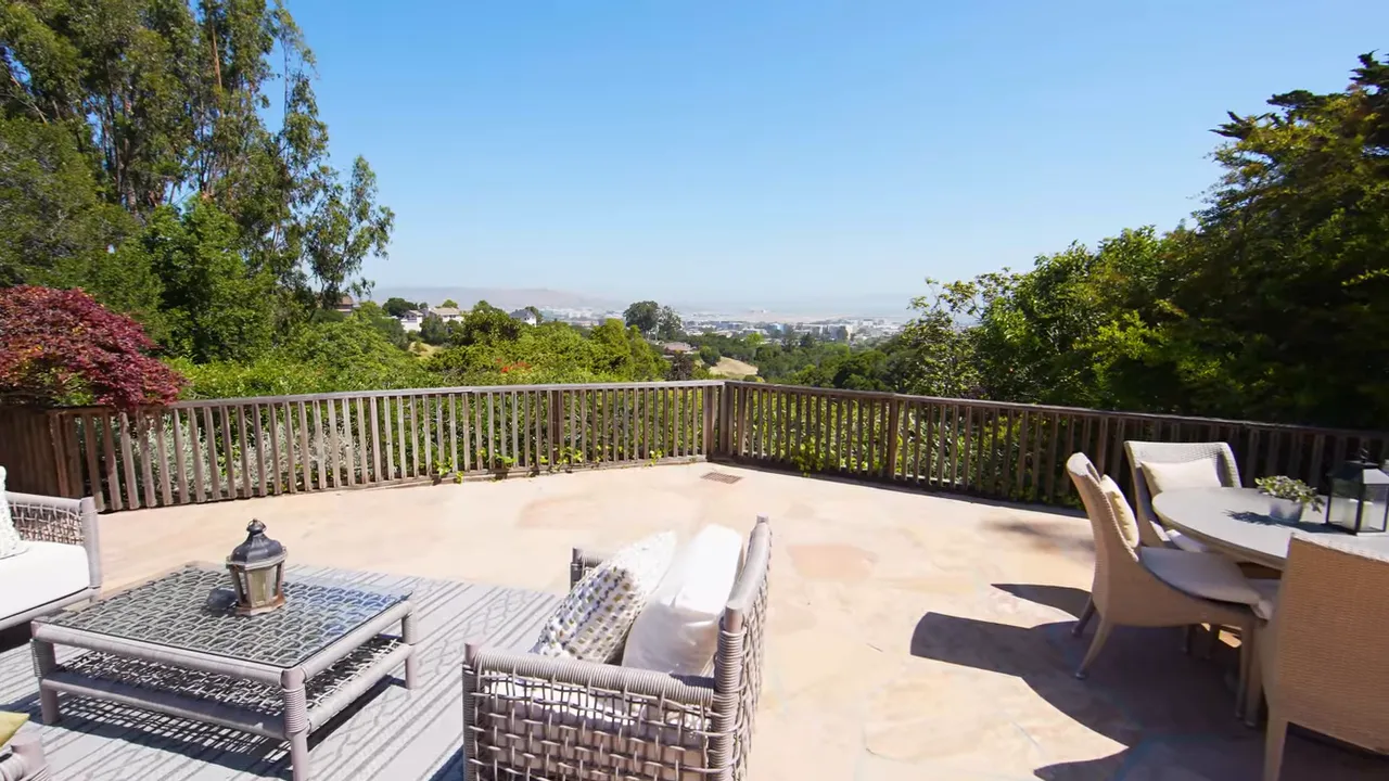 Spacious flat patio with outdoor seating, table and railing overlooking trees and a distant bay view under a clear sky