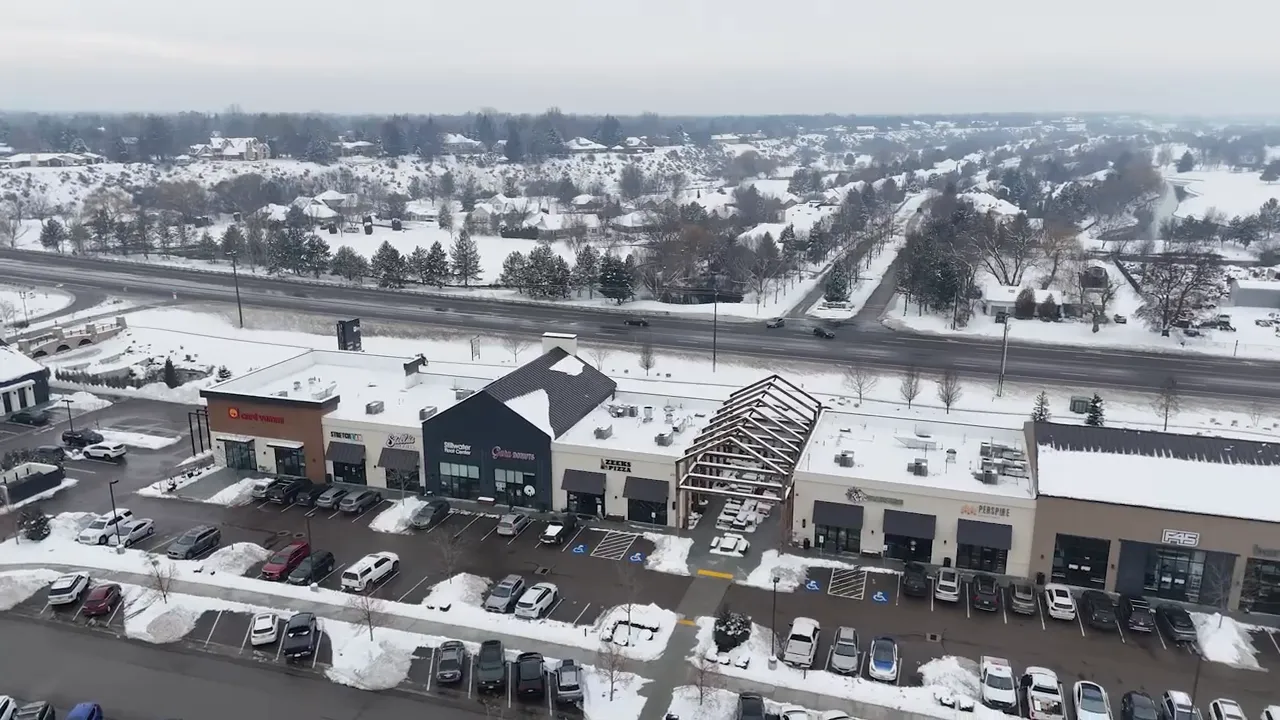 Snowy aerial view of a shopping center with parking lot and surrounding neighborhood