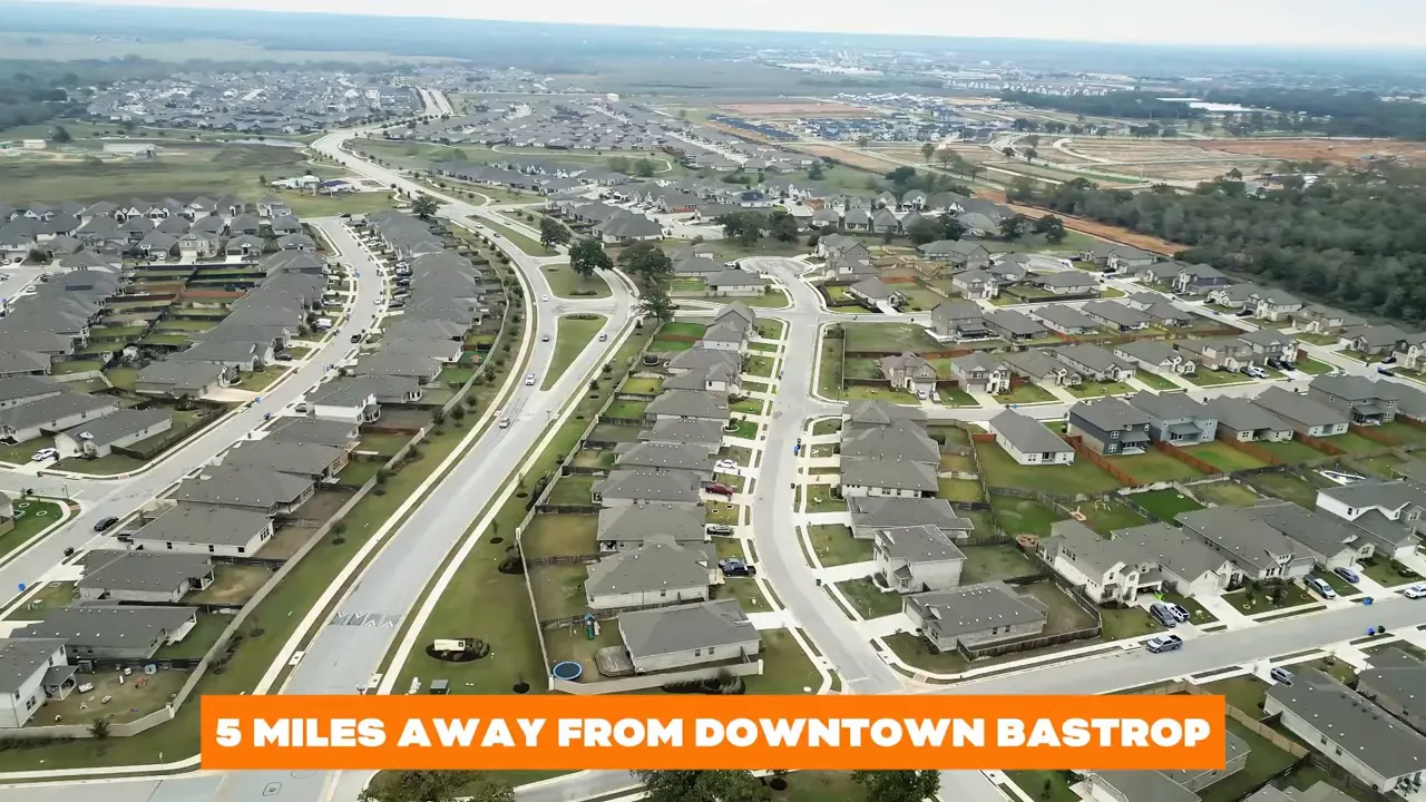 Aerial drone view of The Colony master-plan neighborhood with a banner reading '5 miles away from downtown Bastrop', showing roads and houses